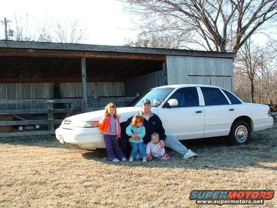 White 1995 Ford Crown Victoria sedan parked on dry grass, with a man and three children posing in front. The car has chrome wheels and tinted windows
