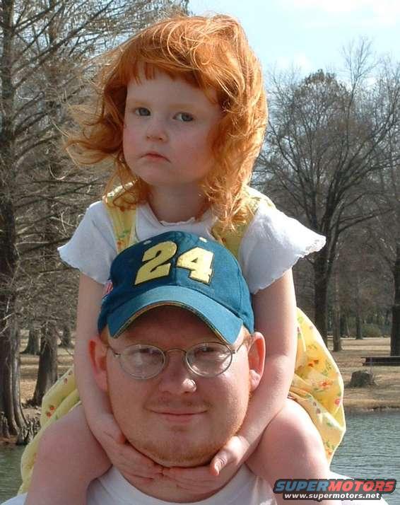 Man wearing glasses and a blue baseball cap with '24' on it, with a young girl with red hair sitting on his shoulders
