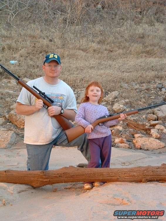 man and young girl holding rifles on a sandy beach with dry brush in background