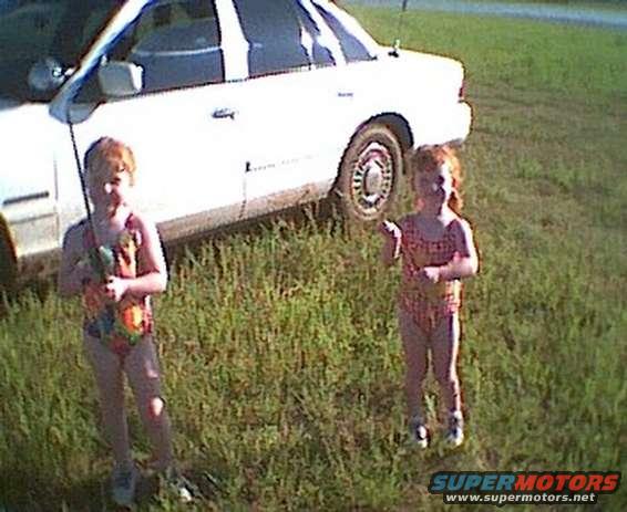 White sedan parked on grass with two young kids in swimsuits standing nearby