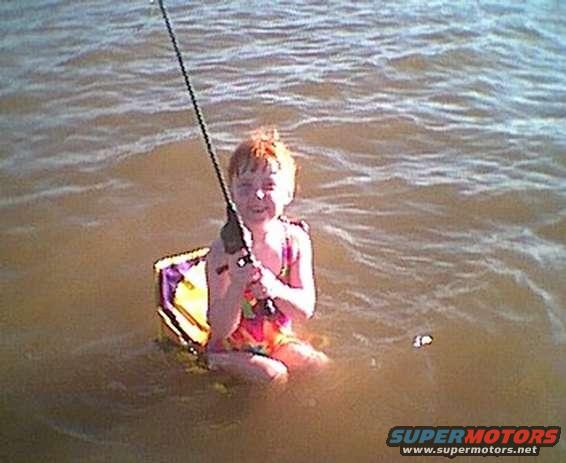 Child with red hair sits in water holding a fishing rod, wearing colorful swim trunks
