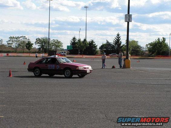 Red 1990 Ford Mustang coupe with racing numbers on side, seen from 3/4 front angle at autocross event
