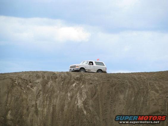 White SUV with a red flag on top, parked on a steep sand dune under a cloudy sky