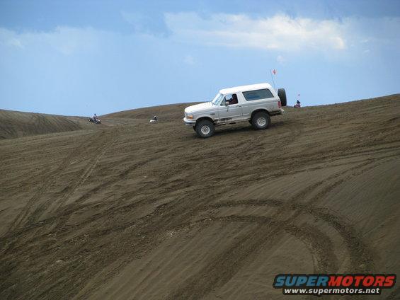 White Ford Bronco driving up a sand dune with a spare tire on the back