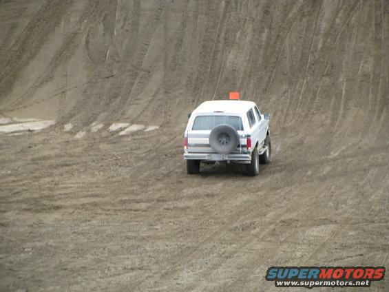 White SUV with spare tire on back driving up a dirt hill. Orange flag on roof