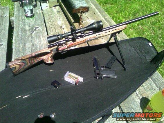 Rifle with scope and bipod, handgun, and ammunition on a black mat