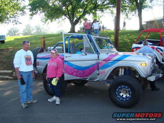 Silver Ford Bronco with pink and blue stripes, roll cage, and off-road tires. Two people stand beside it
