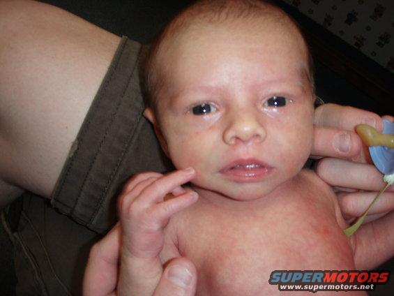 Newborn baby held by adult hands, with a pacifier nearby. Text 'SUPERMOTORS' visible