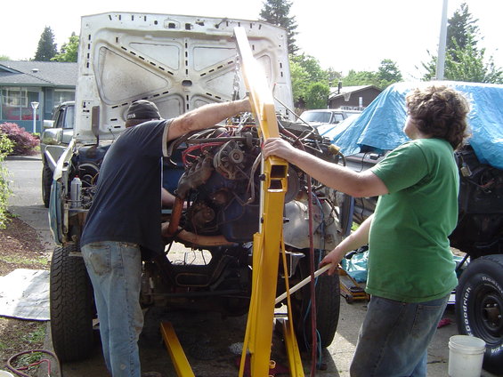 Two guys using an engine hoist to remove the engine from a disassembled vehicle