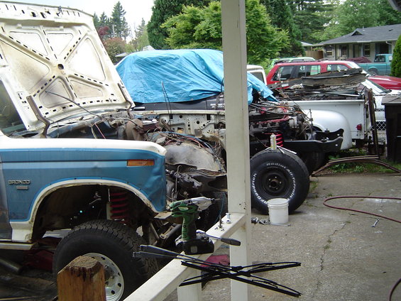 Blue and white Ford Bronco with hood open, undergoing repairs, parked next to a white pickup truck