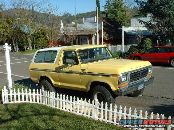 Yellow 1978 Ford Bronco parked on a street with a white picket fence in the foreground