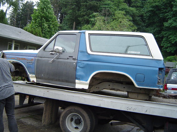Blue and white Ford Bronco body on a flatbed truck, missing doors and wheels