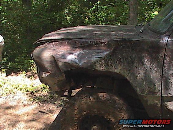 Mud-covered pickup truck with damaged front fender and hood, parked in a wooded area