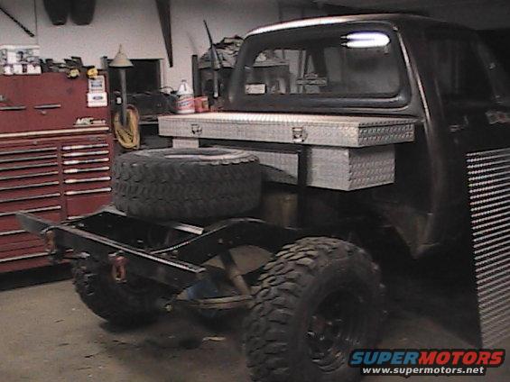Truck chassis with large spare tire, toolbox, and custom rear bed frame. Dark color, appears to be in a garage