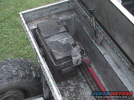 Truck bed with battery box and tire. The battery box is dirty and contains a large battery. A red tool is visible inside the box. The tire is large and off-road style