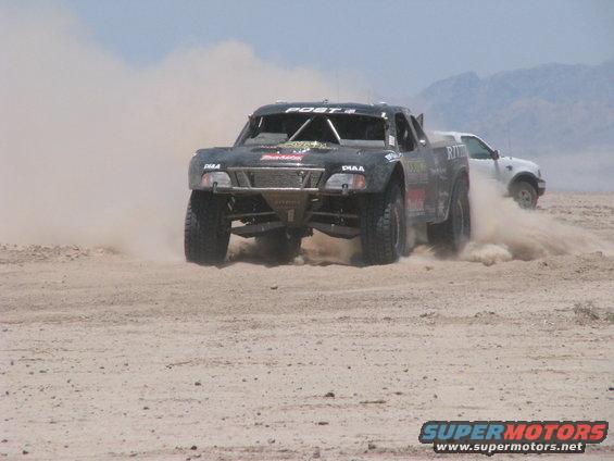 Off-road race truck kicking up dust, another white SUV behind it on a dirt track