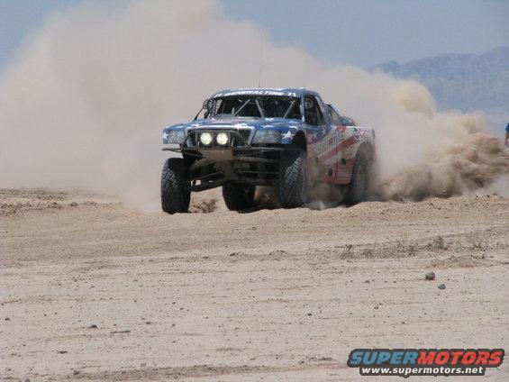 Off-road racing truck with American flag livery kicking up dust, front view
