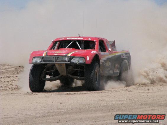 Red off-road racing truck kicking up dust on a dirt track. Number 8 is visible on the door