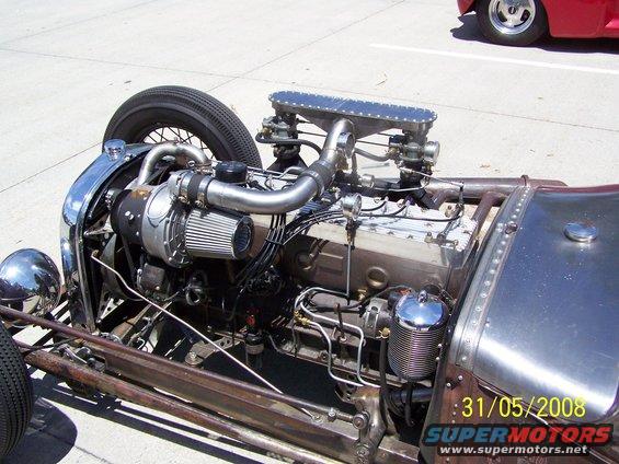 Hot rod engine with turbocharger, chrome accents, and exposed metal bodywork. A red car is partially visible