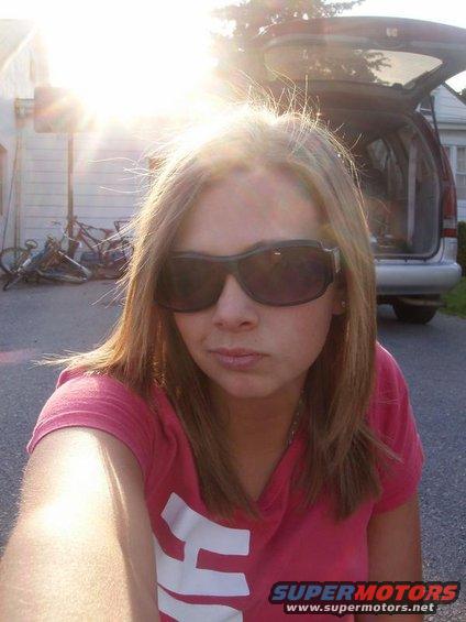 Woman in sunglasses and pink shirt takes a selfie outdoors, with a van and bicycles in the background