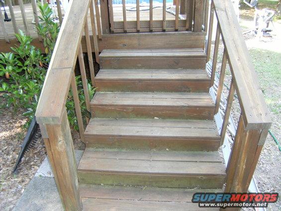 Wooden deck stairs with a railing on the left and right. Greenery is visible to the left of the stairs