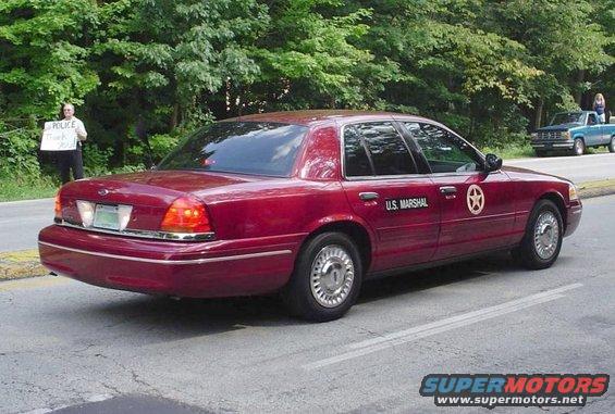 Maroon Ford Crown Victoria U.S. Marshal car parked on a road, with a person holding a 'Police Thank You!' sign in the background