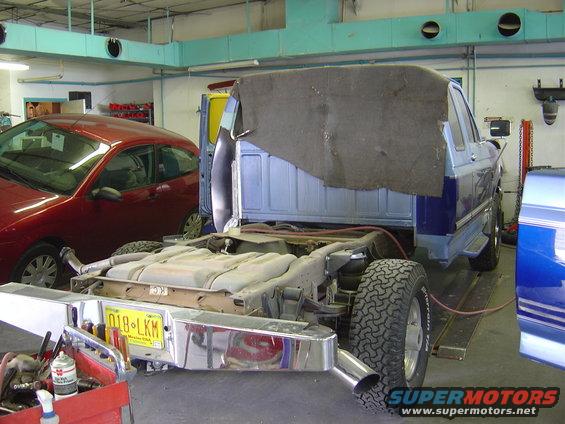 1996 Ford F-250 chassis with bed removed, showing fuel tank and exhaust. Red car in background