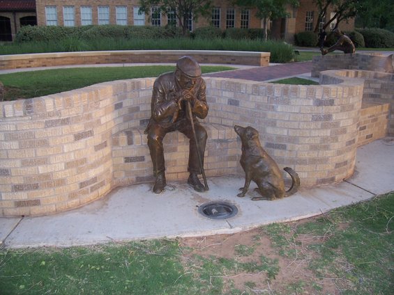 Bronze sculpture of an old man with a cane and a dog sitting on a brick bench outdoors