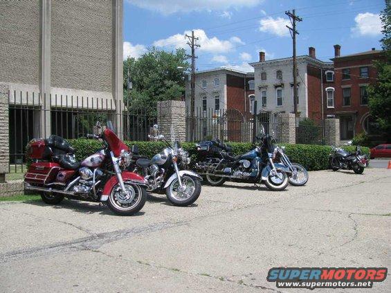 Several motorcycles parked in a lot with buildings in the background