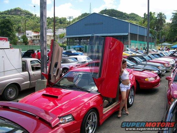 Red sports car with butterfly doors, woman in white shorts standing next to it. Other cars parked in a row behind it