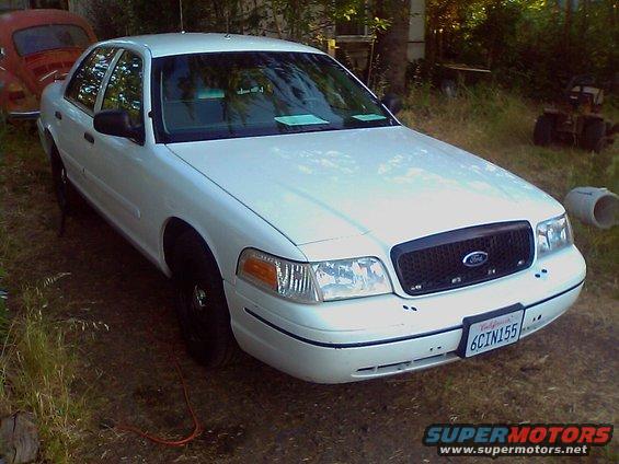 White 2000 Ford Crown Victoria sedan, front 3/4 view, parked outdoors on dirt, with a red car in the background