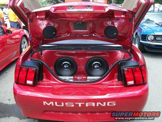 Red 2001 Ford Mustang with trunk open, displaying two large subwoofers and smaller speakers. A blue classic car is visible to the right