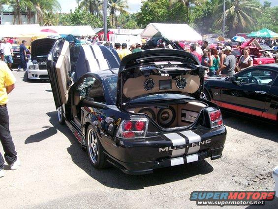 Black Mustang with gullwing doors and trunk open showing speakers. Another Mustang is parked next to it