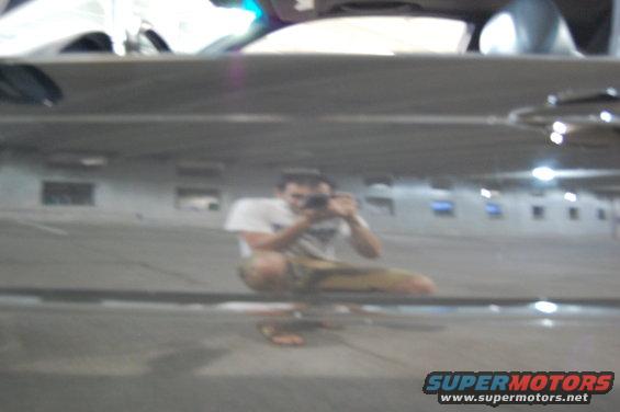 Reflection of a person crouching and taking a photo in a dark, reflective surface, possibly a car