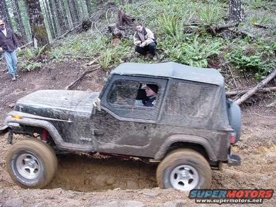 1995 Jeep Wrangler driving through a muddy ditch on a wooded trail