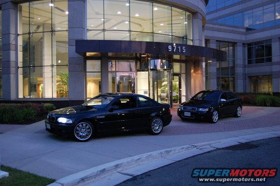 Black 2002 BMW M3 coupe parked in front of a modern building with a black Audi sedan behind it