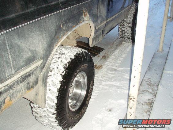 1992 Ford Bronco rear wheel in snow, side view. Mud flaps and large off-road tires are visible