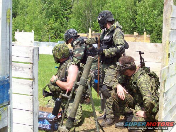paintball players in camouflage gear, one with a mortar, in a wooded area with wooden barriers