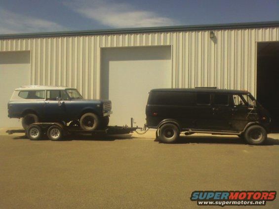 Blue SUV on a trailer, towed by a black van, parked in front of a corrugated metal building