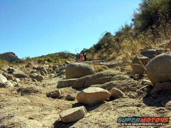 people hiking up a rocky, dry hillside under a clear blue sky. Some large boulders are in the foreground