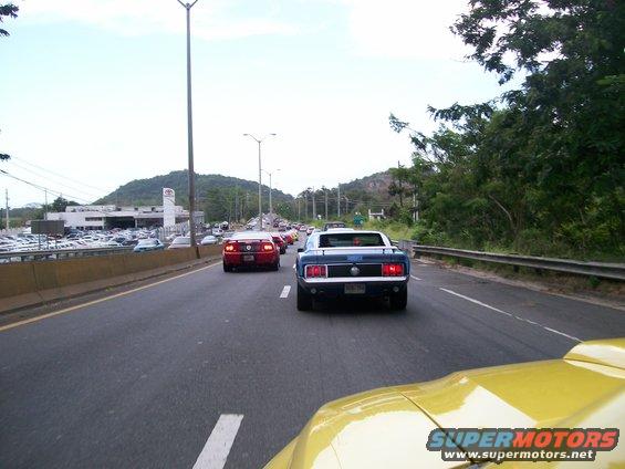 Blue classic car with a black stripe on the trunk, seen from behind on a highway