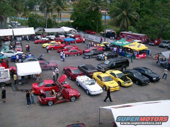 Cars parked at an outdoor event, some with doors open. A white convertible and several red, yellow, and black coupes are visible