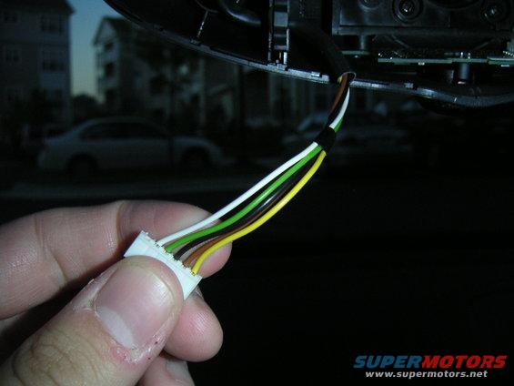 Hand holding a wiring harness with multiple colored wires, with a blurry car and houses in the background