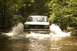 1987 Ford Bronco - A white and green 1987 Ford Bronco splashes through a shallow river surrounded by trees.