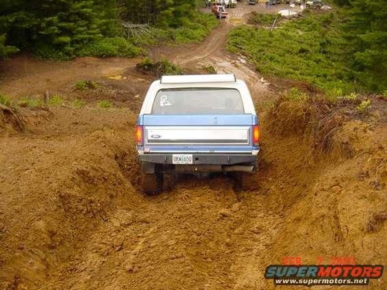 Rear view of a blue and white 1980s Ford Bronco driving on a muddy trail