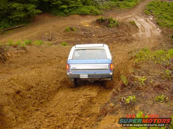 Rear view of a blue and white 1980s Ford Bronco driving on a muddy trail