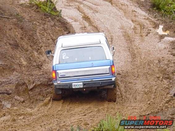 Rear view of a blue and white Ford Bronco driving up a muddy incline. License plate reads OGM650