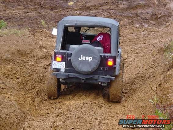 Jeep Wrangler driving on a dirt trail, rear view. It has a soft top and a spare tire on the back