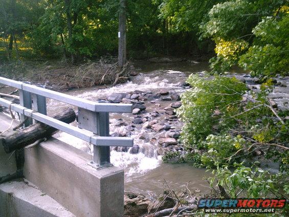 Bridge railing over a fast-moving creek with debris and trees on the banks