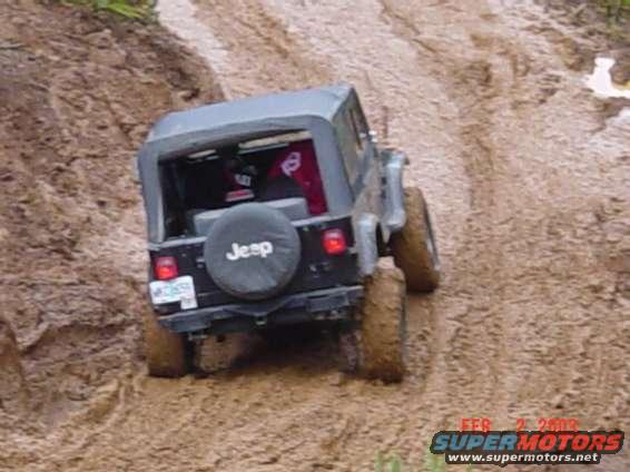 Jeep Wrangler climbing a muddy hill, rear view. Spare tire cover says 'Jeep'